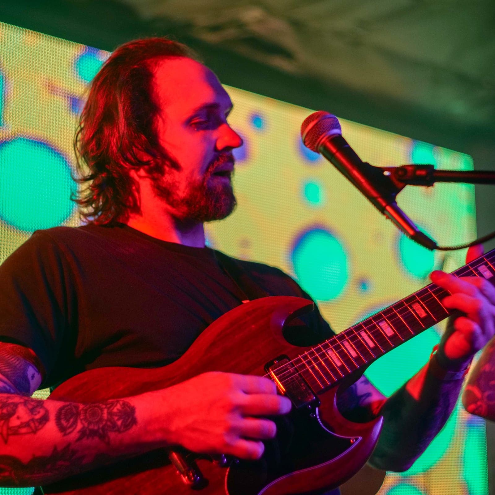 Elliot Preston playing electric guitar beside a microphone under colorful stage lights.